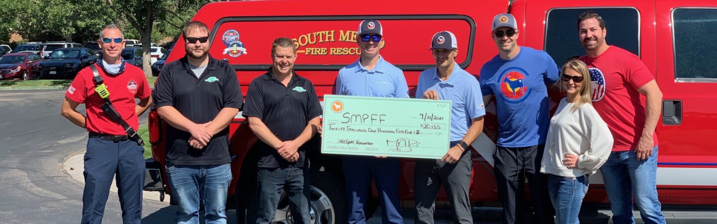 A team of men and women standing in front of a fire truck holding a large fundraiser check